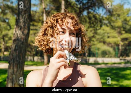 Nahaufnahme einer jungen, sportlichen Rothaarigen Frau, die einen Sport-BH trägt, während sie einen Ernährungsstab beißt. Athletische Frau, die einen Proteinriegel isst. Schöner Fitnessbereich Stockfoto