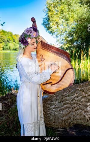 Die junge ukrainische Frau in Nationaltracht spielt an einem sonnigen Sommertag eine Bandura am Fluss. Stockfoto