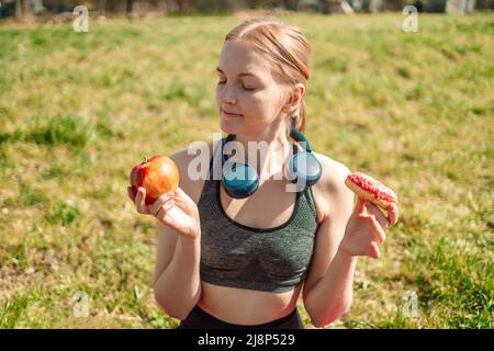 Auswahl zwischen Apfel und Donut. Diät-Konzept. Blonde Mädchen hält einen rosa Donut und Apfel. Süßigkeiten sind ungesunde Junk Food. Diät. Gesunde Ernährung. Stockfoto