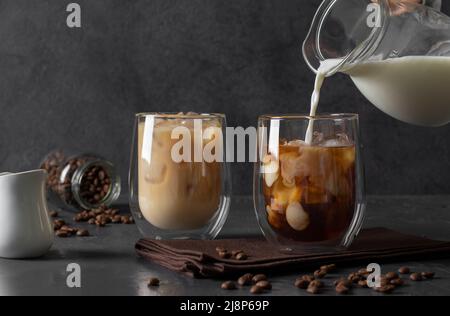 Zwei Gläser kalten Kaffees mit Eiswürfeln, in die Milch auf dunklem Grund gegossen wird Stockfoto