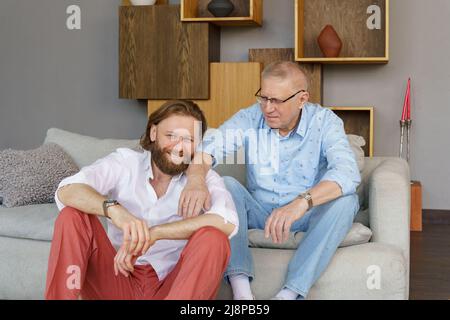 Vater und Sohn sitzen auf der Couch im Wohnzimmer, beide Männer schauen in die Kamera, ein älterer Mann in Brille mit Dioptrien, ein stilvoller junger Mann mit einem Stockfoto