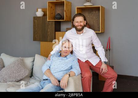Vater und Sohn sitzen auf der Couch im Wohnzimmer, beide Männer schauen in die Kamera, ein älterer Mann in Brille mit Dioptrien, ein stilvoller junger Mann mit einem Stockfoto
