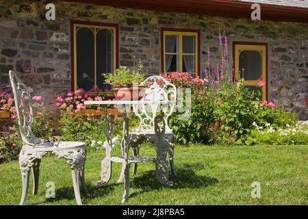 Weiß gusseiserne viktorianische Gartenstühle und Tisch auf dem vorderen Rasen des alten, ca. 1794 französischen Regimehüttenstil Feldsteinhauses im Sommer. Stockfoto