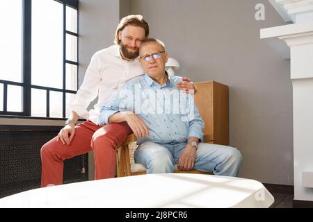 Vater und Sohn sitzen auf dem Stuhl im Wohnzimmer, beide Männer blicken in die Kamera, ein älterer Mann in einer Brille mit Dioptrien, ein stilvoller junger Mann mit einem Stockfoto