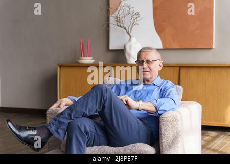 Ein älterer Mann in einer Brille mit Dioptrien sitzt auf der Couch im Wohnzimmer, er schaut in die Kamera Stockfoto