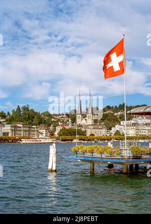 Nationalflagge der Schweiz, die am sonnigen Herbsttag über den Vierwaldstättersee fliegt, mit den Zwillingstürmen von St. Leodegar im Hintergrund, Stockfoto