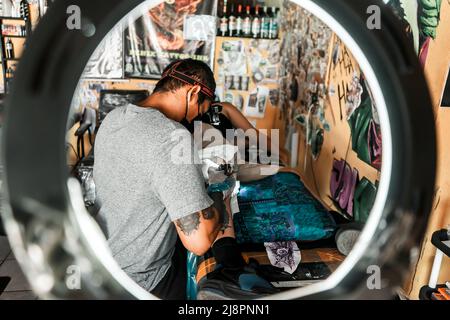 Latino-Tattoo-Künstler arbeitet an einem Design, das von einem Lichtring in seinem Atelier aus gesehen wird Stockfoto