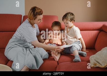 Mutter liest Buch mit zwei Kindern auf dem Sofa. Glückliche Familienmomente zu Hause. Zeit verbringen und Kinder erziehen. Geschwister mit Mama spielen, beobachten Stockfoto