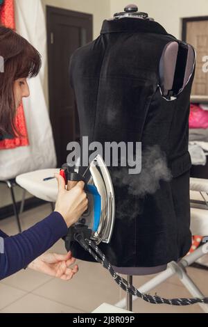 Eine Näherin bügelt eine unvollendete Jacke auf eine Schaufensterpuppe. Stockfoto