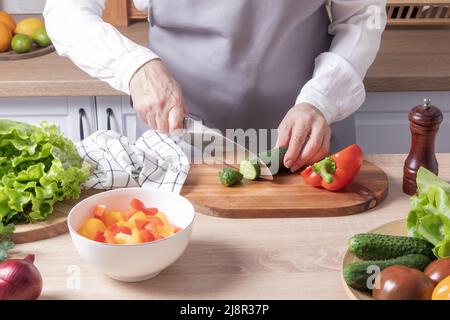 Eine ältere Frau in einer grauen Schürze schneidet eine frische grüne Gurke auf einem Schneidebrett in einer modernen Küche. Zubereitung von Gemüsesalat Stockfoto