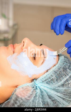Die Kosmetikerin mit Pinsel trägt eine photochemische und glykolische Peeling-Gesichtsmaske auf das weibliche Gesicht der Patientin in der Schönheitsklinik auf Stockfoto