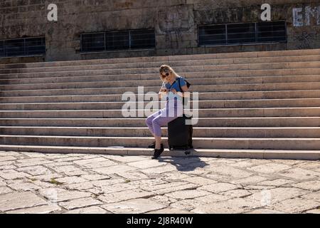 Eine blonde Frau mit Sonnenbrille sitzt auf dem Gepäck auf einer großen Treppe in der Nähe eines Steingebäudes. Suche nach elektronischen Tickets und Karten Stockfoto