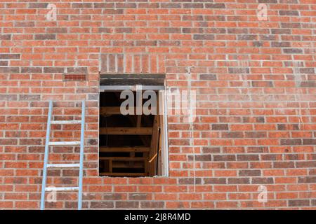 Eine Aluminiumleiter, die sich an einer roten Ziegelwand lehnt und zu einer leeren Fensteröffnung eines neu erbauten Hauses führt Stockfoto