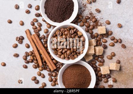 Draufsicht auf geröstete Bohnen mit gemahlenem Kaffee in weißen Schüsseln und braunem Zucker, Zimtstangen auf grauem Betongrund Stockfoto