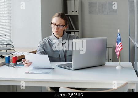 Ernsthafte weibliche Visumarbeiterin, die eine Brille trägt, sitzt am Schreibtisch im Büro und schaut sich die Unterlagen des Antragsformulars an Stockfoto