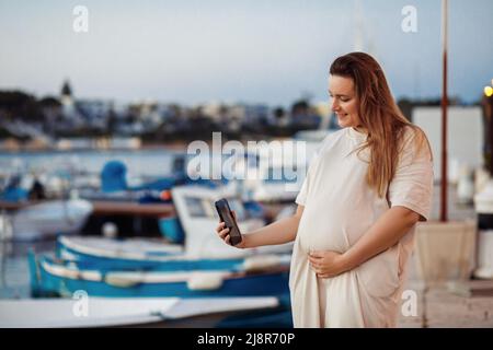 Fröhliche und ruhige blonde langhaarige Schwangere lächelt, schaut auf den Bauch, berührt den Bauch, nimmt Selfies und Videoanruf Stockfoto