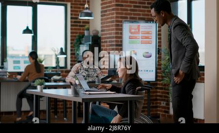 Junger Mann hilft Geschäftsfrau im Rollstuhl, am Schreibtisch zu arbeiten und unterstützt sie im Büro. Weibliche Arbeitnehmer mit Behinderung Planung Führungsanalyse mit finanziellen Daten Charts und Diagramm. Stockfoto