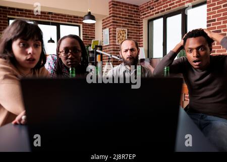 Schockierte multiethnische Freunde, die zu Hause tragische Nachrichten auf einem Laptop sahen. Ängstliche, vielfältige Menschen, die online Horrorfilme im Internet anschauen, während sie am Tisch im Wohnzimmer sitzen. Stockfoto