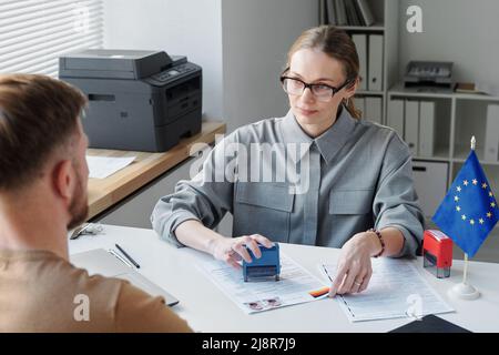 Moderner Konsularbeamter, der am Tisch sitzt und das Interview mit einem unkenntlichen Mann beendet, der den Visumantrag genehmigt Stockfoto