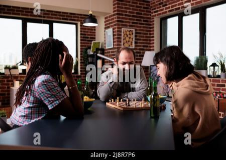 Multiethnische Gruppe von Freunden, die zu Hause Schach spielen. Verschiedene Menschen sitzen zu Hause, im Wohnzimmer, genießen Brettspiele, Snacks und Getränke, während sie eine gute Zeit zusammen haben. Stockfoto