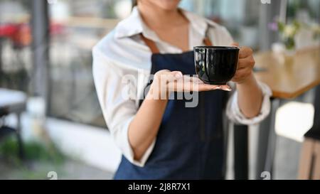 Zugeschnittenes Bild, weibliche Barista hält eine schwarze Tasse Kaffee oder einen Kaffeebecher in ihrem Café. Stockfoto