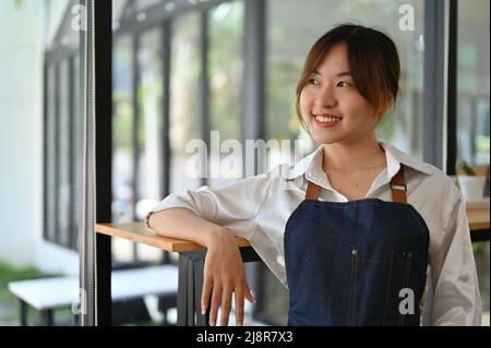 Porträt eines erfolgreichen asiatischen Jungunternehmers, Cafés oder Café-Besitzers, lächelnd und mit Leidenschaft aus dem Fenster blickend. Stockfoto