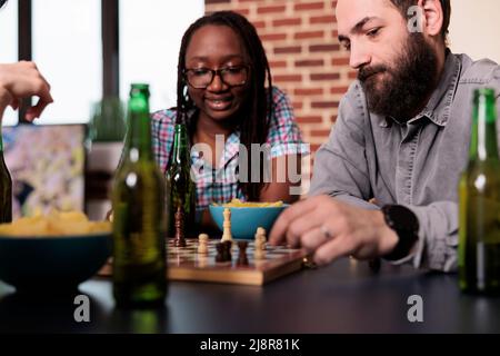 Intelligente multiethnische Freunde sitzen zu Hause, während sie gemeinsam Schach spielen. Verschiedene Menschen im Wohnzimmer entspannen sich bei einem strategischen Brettspiel, während sie Snacks und Getränke genießen. Stockfoto