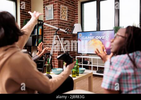 Verärgerte Frau reagiert auf Spiel über Bildschirm, während Videospielrunde auf moderner Konsole zu verlieren. Verschiedene Spieler verärgern sich über verlorene Spiele, während sie mit engen Freunden auf dem Sofa im Wohnzimmer sitzen. Stockfoto