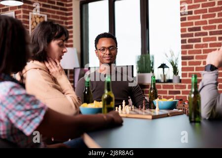 Lächelnder Mann, der mit Freunden am Tisch im Wohnzimmer sitzt, während er eine Partie Schach beobachtet. Intelligente, multiethnische Menschen spielen Strategiespiele und genießen dabei Snacks und Getränke. Stockfoto