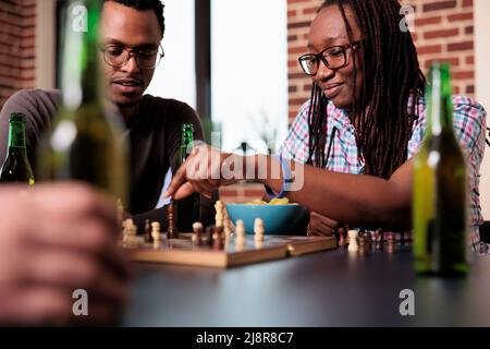 Eine kluge Frau, die ein Schachstück an Bord bewegt, während ihre Freundin neben ihr sitzt. Intelligente junge Menschen zu Hause im Wohnzimmer spielen strategische Brettspiele, während sie Snacks und Getränke genießen. Stockfoto