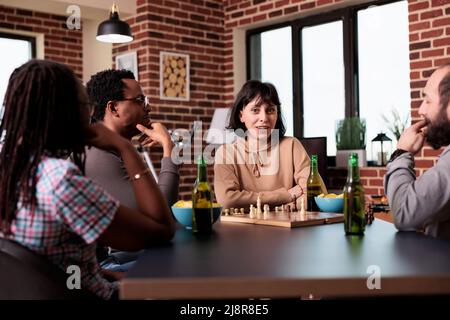 Lächelnde Frau, die im Wohnzimmer am Tisch sitzt, mit multiethnischen Menschen, die Schach spielen. Clevere, vielseitige Freunde genießen gemeinsam Strategiespiele und haben Spaß mit Snacks und Getränken. Stockfoto