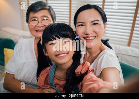 Nettes asiatisches Mädchen lächelt und schaut in die Kamera, während sie Selfie mit Großmutter und Tante nimmt, während sie im Wohnzimmer zu Hause auf dem Sofa sitzt Stockfoto