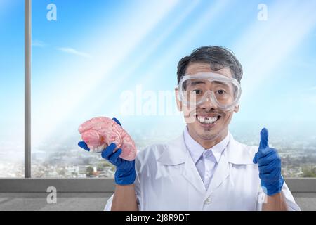 Asiatischer Nerd-Wissenschaftler steht und zeigt den Daumen hoch, während er ein menschliches Gehirn im Labor hält Stockfoto