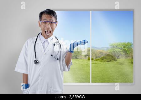 Asiatischer Nerd-Wissenschaftler mit einem Stethoskop, das steht und etwas mit einem Fensterglashintergrund zeigt Stockfoto