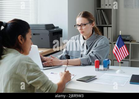 Eine hispanische Frau sitzt an der Rezeption des US-Botschaftsbüros und unterschreibt das Visaantragsformular, während ein Konsularbeamter die Informationen auf dem Laptop überprüft Stockfoto