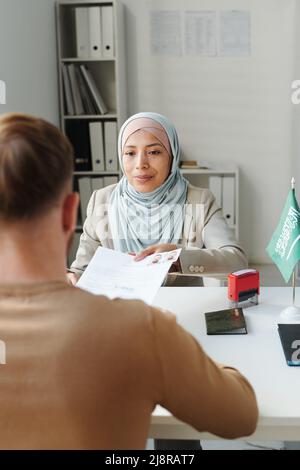 Eine moderne muslimische Frau, die in der Botschaft arbeitet, sitzt an der Rezeption vor einem Mann, der ihm nach der Genehmigung des Visums Dokumente übergibt Stockfoto