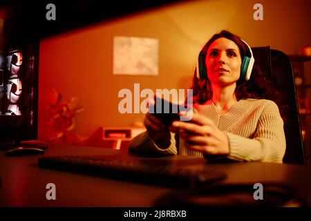 Junge Erwachsene kaukasische Frau mit langen lockigen Haaren, die Kopfhörer trägt, genießt es, nachts Videospiele zu spielen Stockfoto