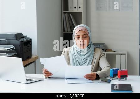 Moderne muslimische Frau mit Hijab sitzt am Schreibtisch im Büro und macht Schreibarbeit, um die Richtigkeit der Informationen zu überprüfen Stockfoto