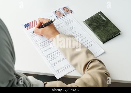 Man kann die nicht erkennbare muslimische Frau, die am Schreibtisch sitzt, mit einem Reisepass und dem Ausfüllen des Visaantrags sehen Stockfoto