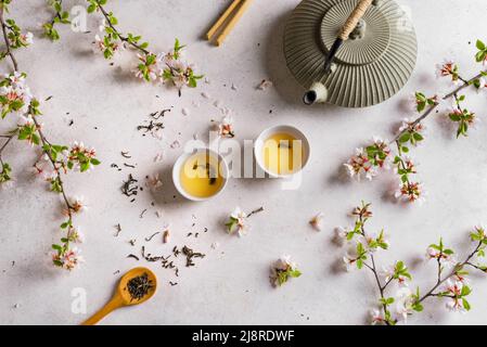 Grüner Tee und Kirschblüte auf weißem Steintisch. Japanische Teekannen aus Gusseisen und Tassen, asiatische Grüntee-Komposition mit Sakura-Blüte und Blütenblättern. Stockfoto