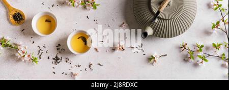 Grüner Tee und Kirschblüte auf weißem Tisch, Banner. Japanische Teekannen aus Gusseisen und Tassen, asiatische Grüntee-Komposition mit Sakura-Blüte und Blütenblättern. Stockfoto