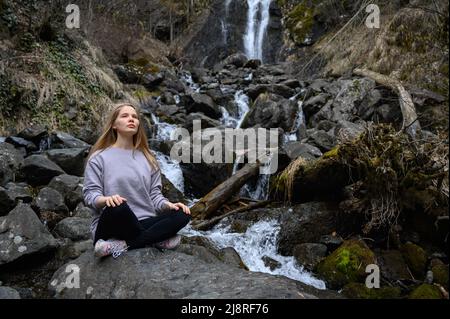 Mädchen sitzt auf einem großen Stein und meditiert in der Nähe eines Wasserfalls neben einem Bergfluss. Stockfoto