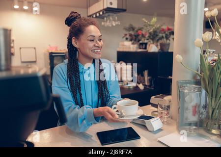 Junge, hübsche Barista mit gemischter Rasse, die dem Gast eine Tasse Kaffee überreicht. Barkeeper arbeiten. Stockfoto