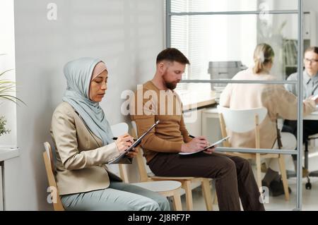 Kaukasischer Mann und muslimische Frau im Hijab sitzen auf Stühlen in der Botschaft oder Visa-Service-Agentur Ausfüllen Antragsformulare, während in der Warteschlange warten Stockfoto