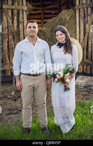 Liebevoller Bräutigam in einer schwarzen Strickjacke und eine niedliche Braut in einem teuren Kleid umarmen sich, sitzen im Wald auf der Natur. Hochzeits-Porträt von Lächeln Stockfoto