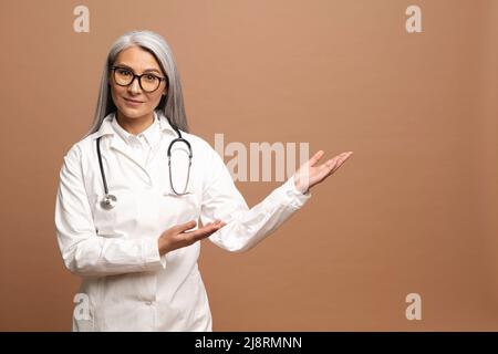 Asiatische charmante Ärztin Pädiatrie, Arzt, Therapeut trägt ein weißes medizinisches Kleid mit einem Stethoskop auf den Schultern zeigt die Hände auf leeren Raum, Mock-up, Kopierer Raum. Gesundheitswesen und Medizin Stockfoto