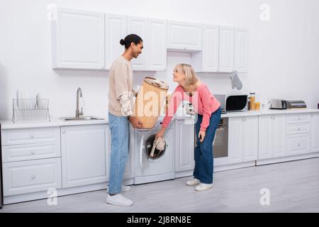 Glückliche blonde Frau, die in der Nähe von afroamerikanischem Mann Kleidung in die Waschmaschine legt Stockfoto