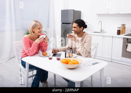 Glückliches multiethnisches Paar, das beim Frühstück Tee trinkt und sich anschaut Stockfoto