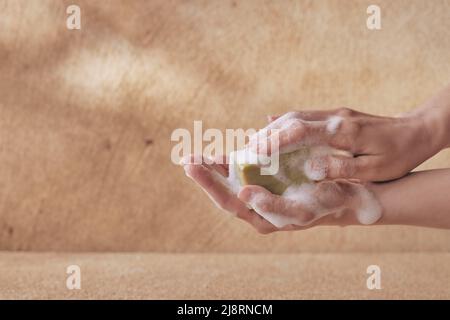 Crop weibliche Hand hält Olivenseife mit Schaum mit Copyspace auf braunem Hintergrund für Gesundheits- und Waschkonzepte Stockfoto