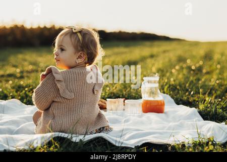 Rückansicht des kleinen Mädchens, das auf einem Plaid im Freien bei Sonnenuntergang sitzt Stockfoto